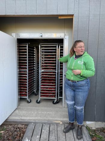 A person standing next to the open door to a building, showing wheeled racks of red peppers inside.